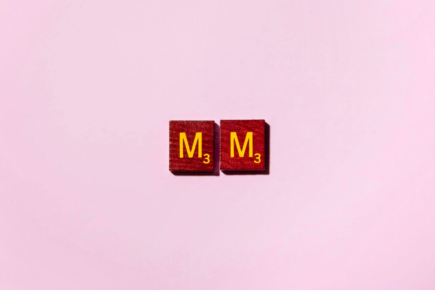 Two Scrabble tiles displaying the letter "M" with a value of 3, positioned on a light pink surface.