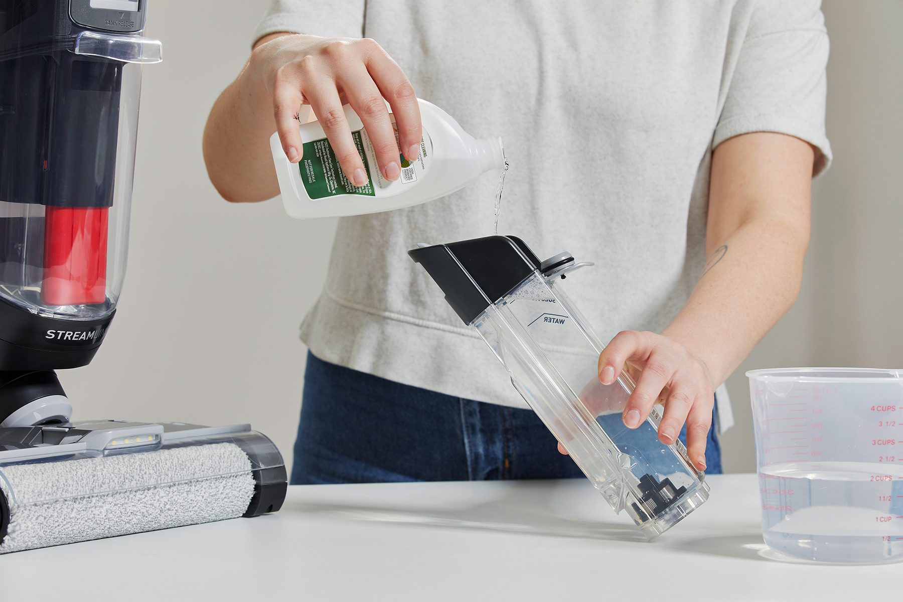 Person pours liquid into vacuum's water container, standing on table with measuring cup and vacuum cleaner nearby.