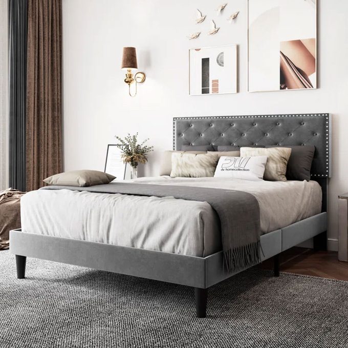A gray upholstered bed stands in a modern bedroom, adorned with decorative pillows, soft lighting, and abstract wall art.