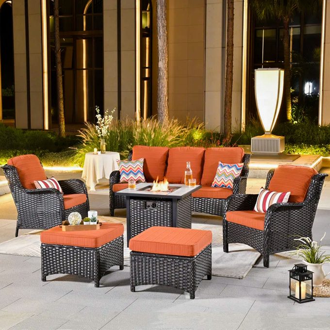 Wicker patio furniture with orange cushions surrounds a lit fire table. Lanterns and plants adorn the modern, illuminated building backdrop.