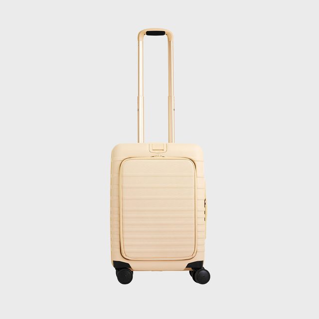 Suitcase stands upright, with telescopic handle extended, against a plain background. It has a beige color and four wheels for mobility.