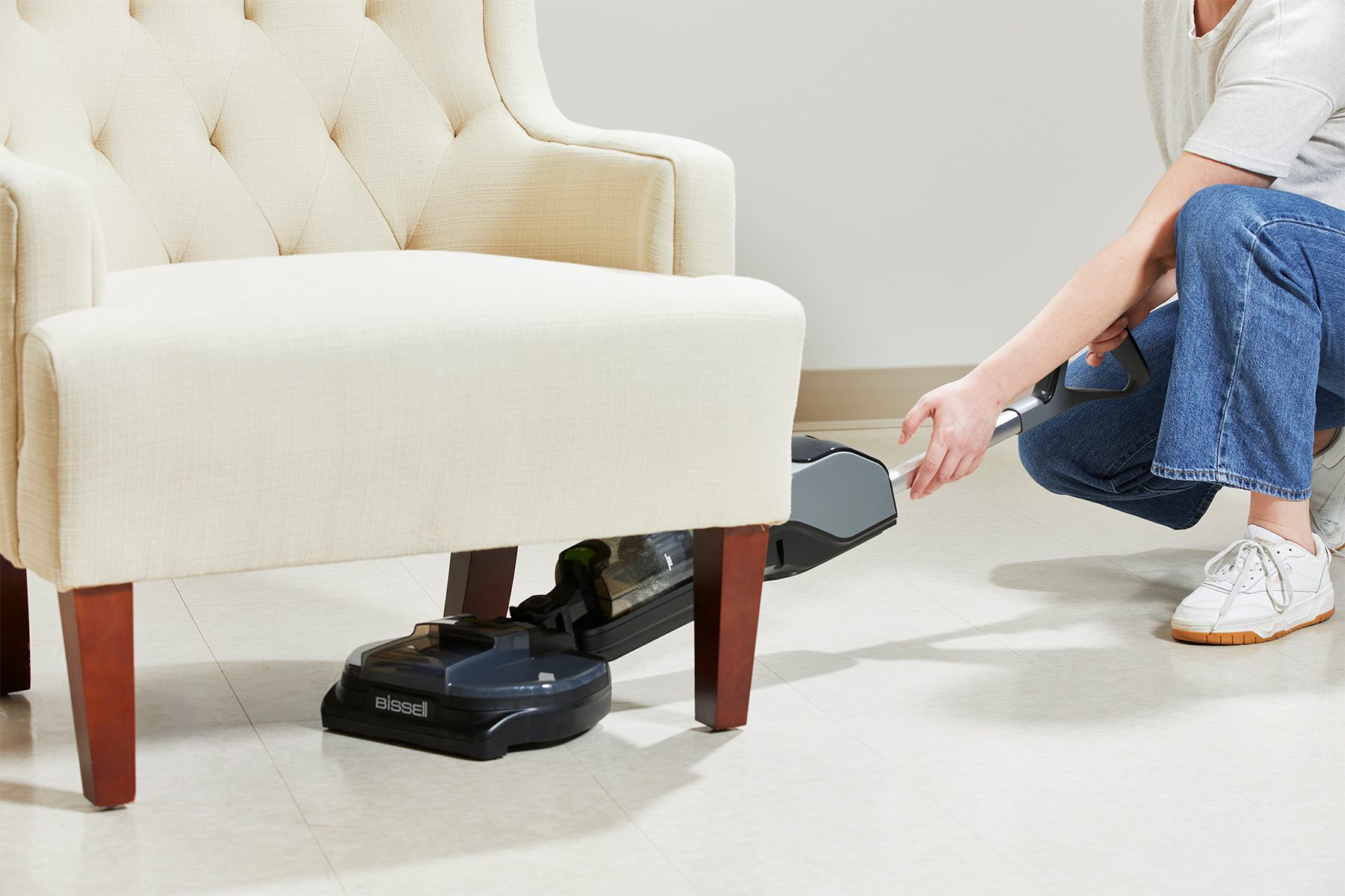 Vacuum cleaner sweeps under beige chair, handled by crouching person wearing jeans and white sneakers on a tiled floor.