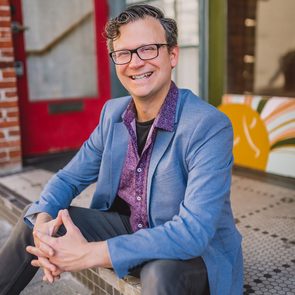 Person sitting smiling, wearing glasses and a blue blazer, in front of a red door and mosaic-tiled area.