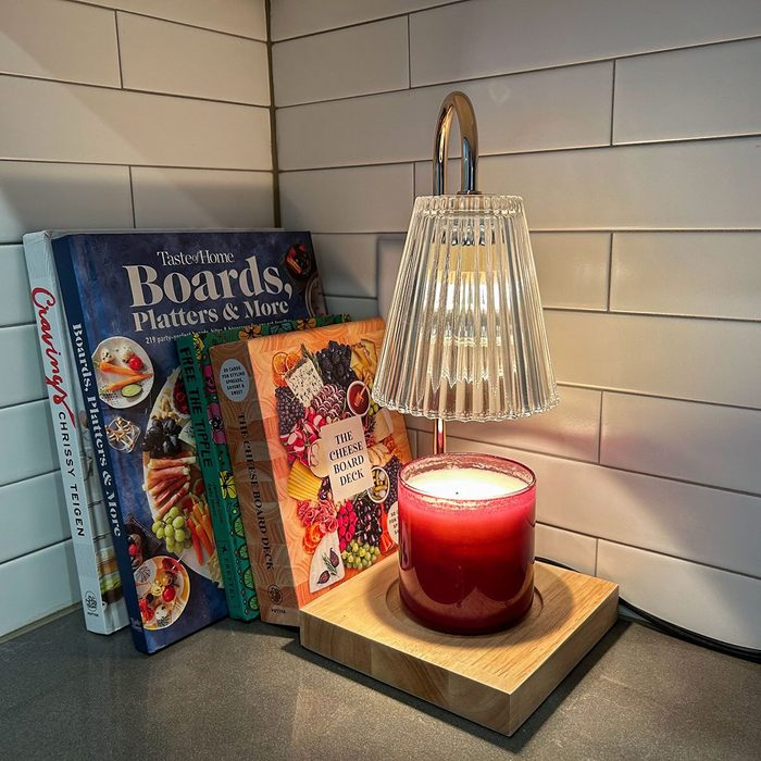 A glass lamp illuminates a red candle on a wooden base beside cookbooks in a tiled kitchen corner.