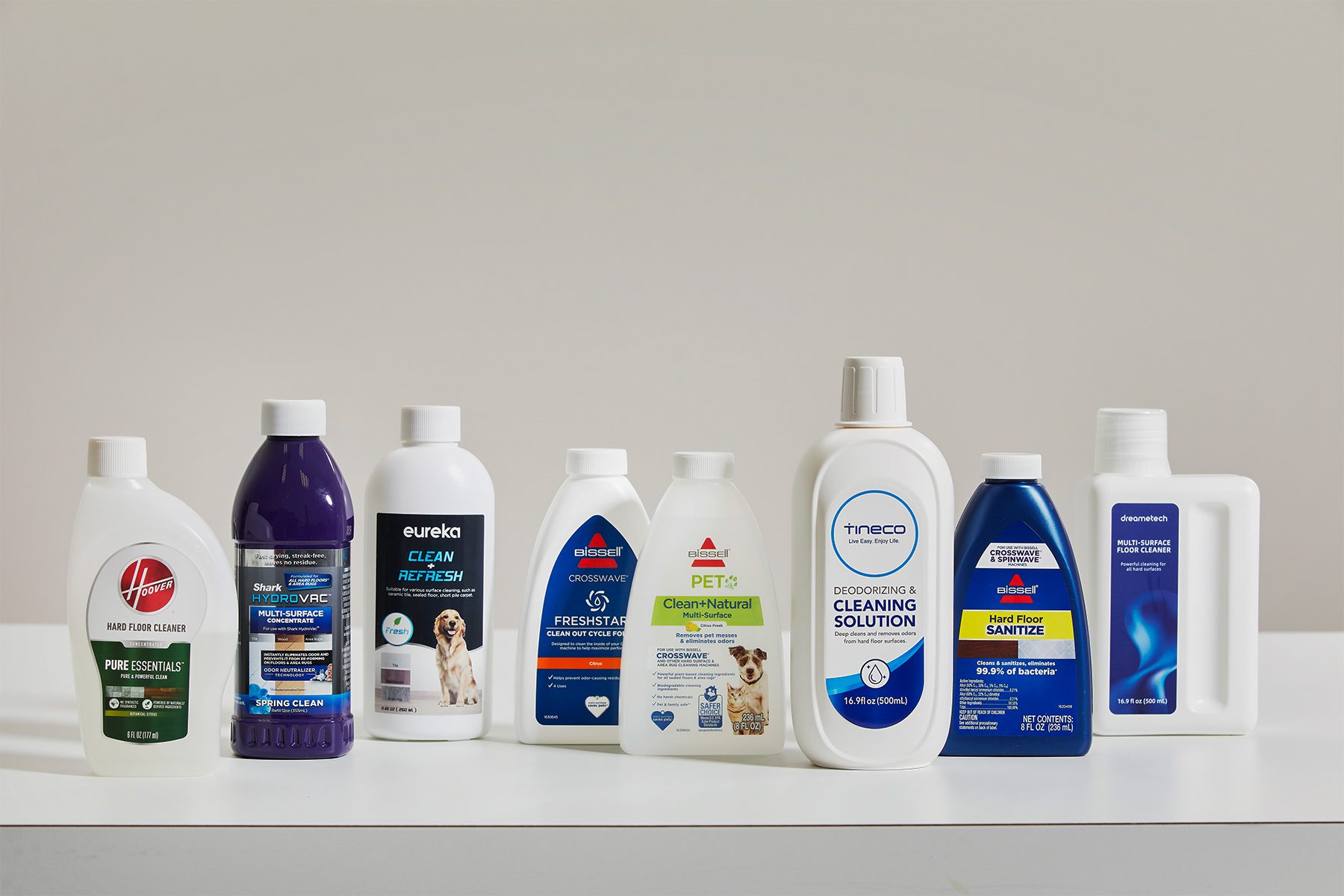 Bottles of floor cleaners are lined up on a white surface, displaying various brands and labels in a well-lit, plain indoor setting.