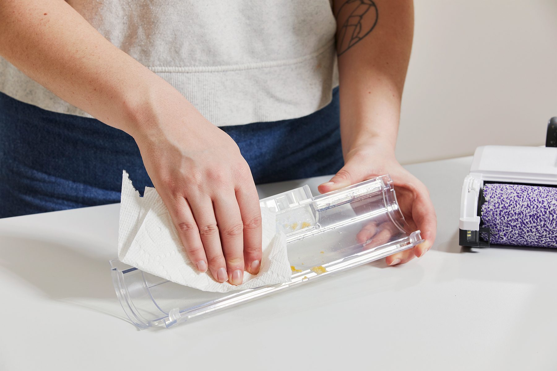 Hands clean a transparent vacuum part using a paper towel, placed on a table with a nearby cleaning device.