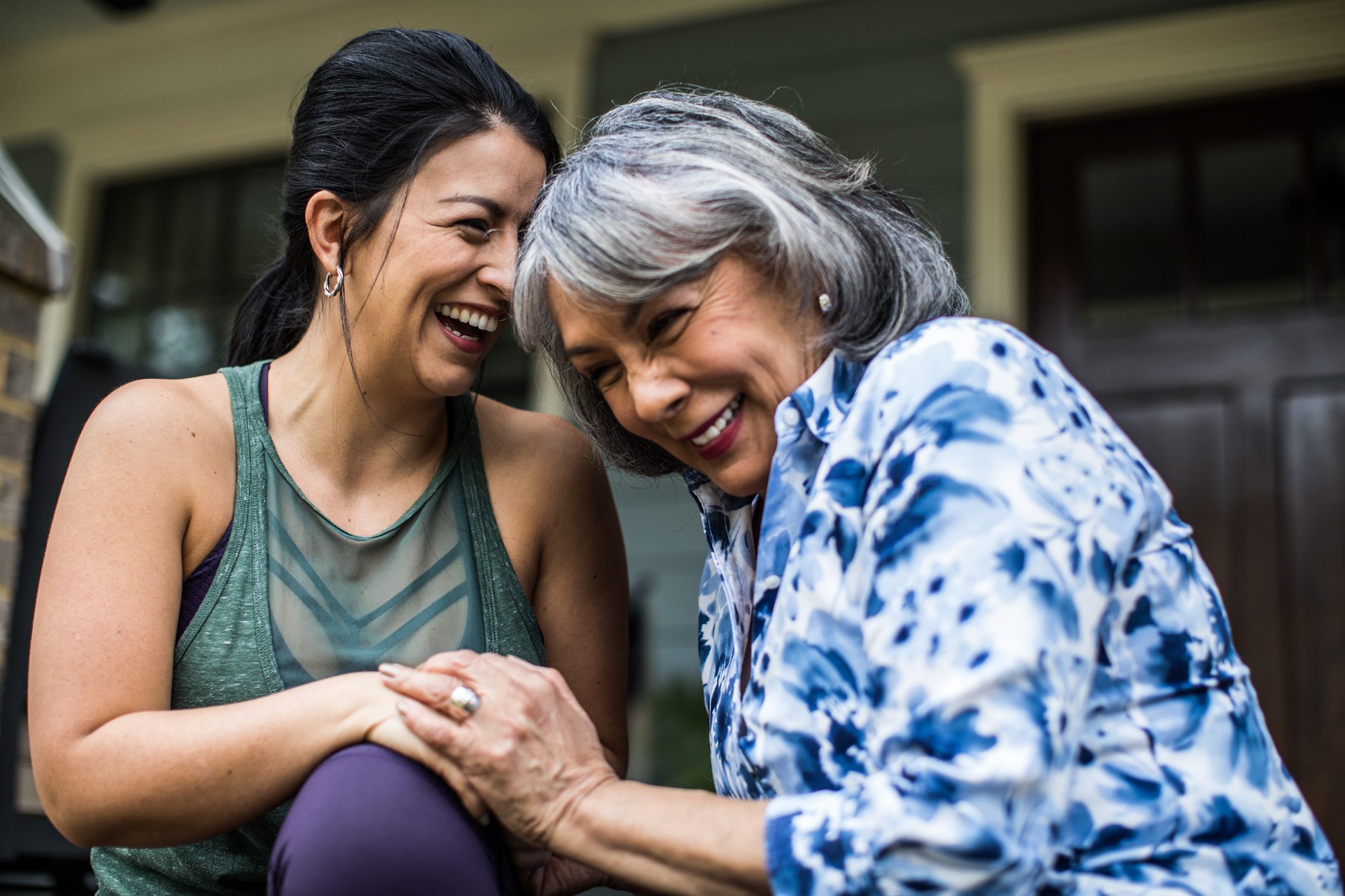 Two women laughing together on a porch, one wearing a green top, the other in a blue patterned shirt.