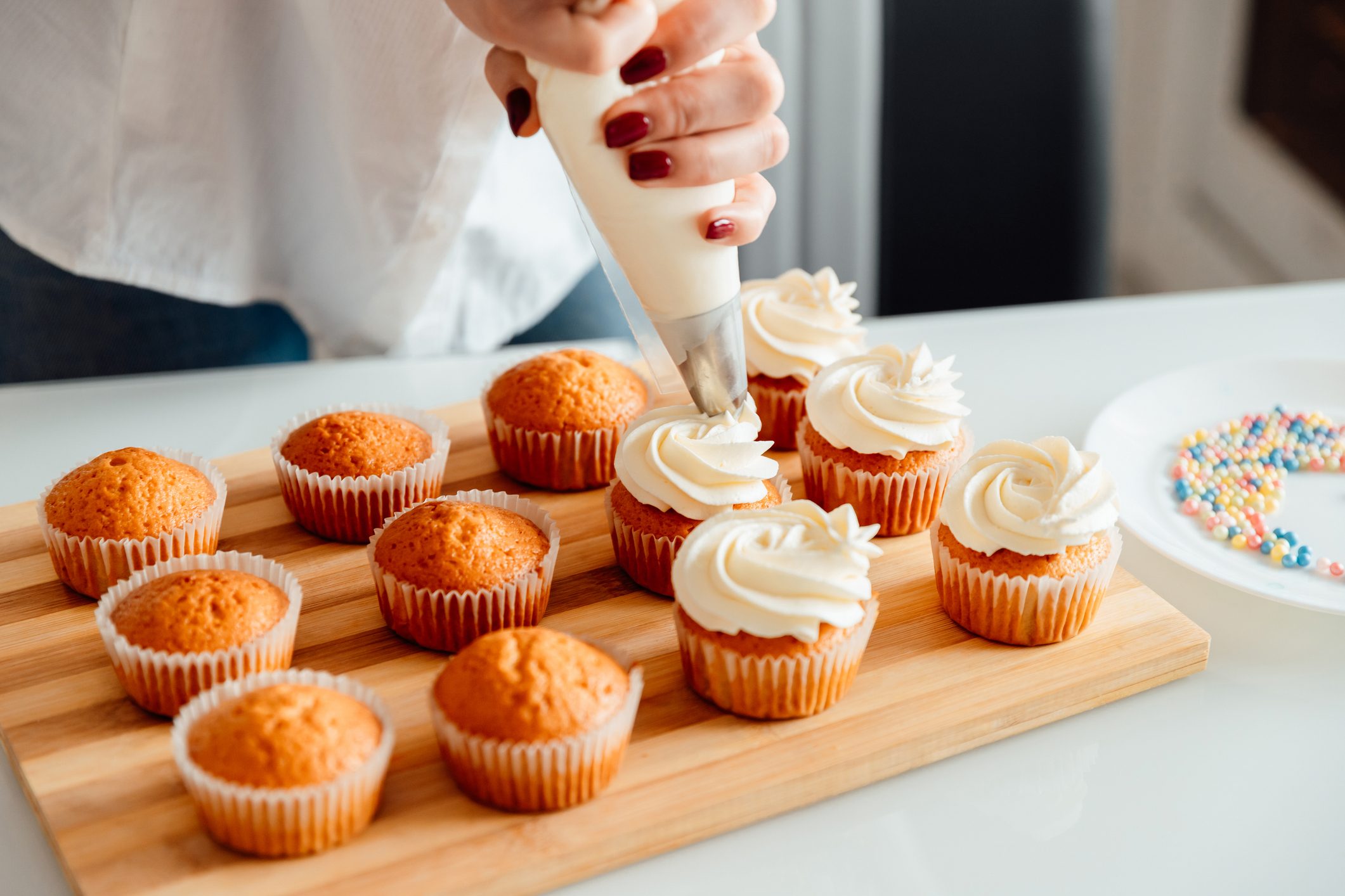 Hand pipes frosting onto cupcakes arranged on a wooden board; colorful sprinkles sit on a nearby white plate.