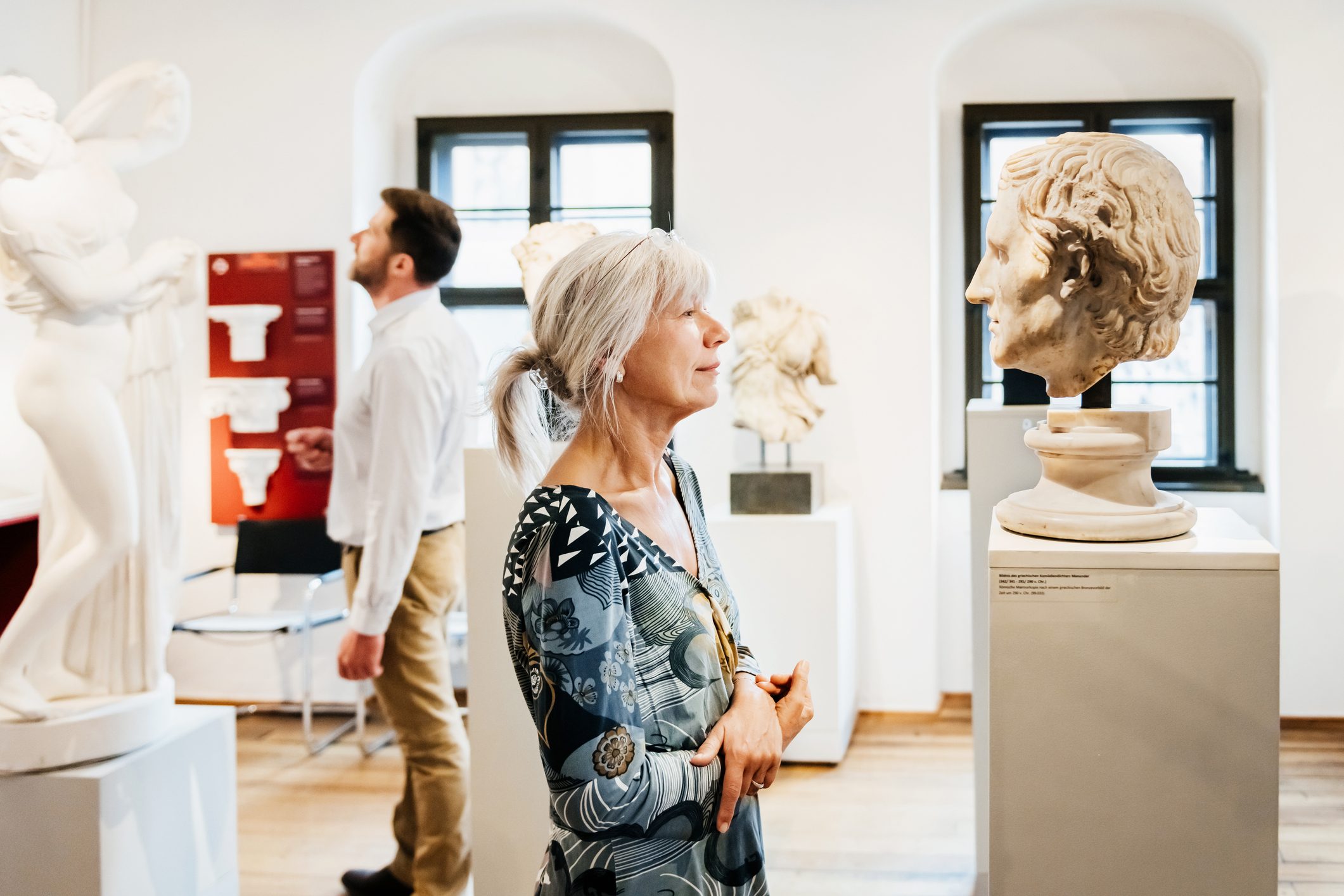 A woman observes a classical sculpture in a museum; a man examines another statue nearby, surrounded by bright, white walls and statues.