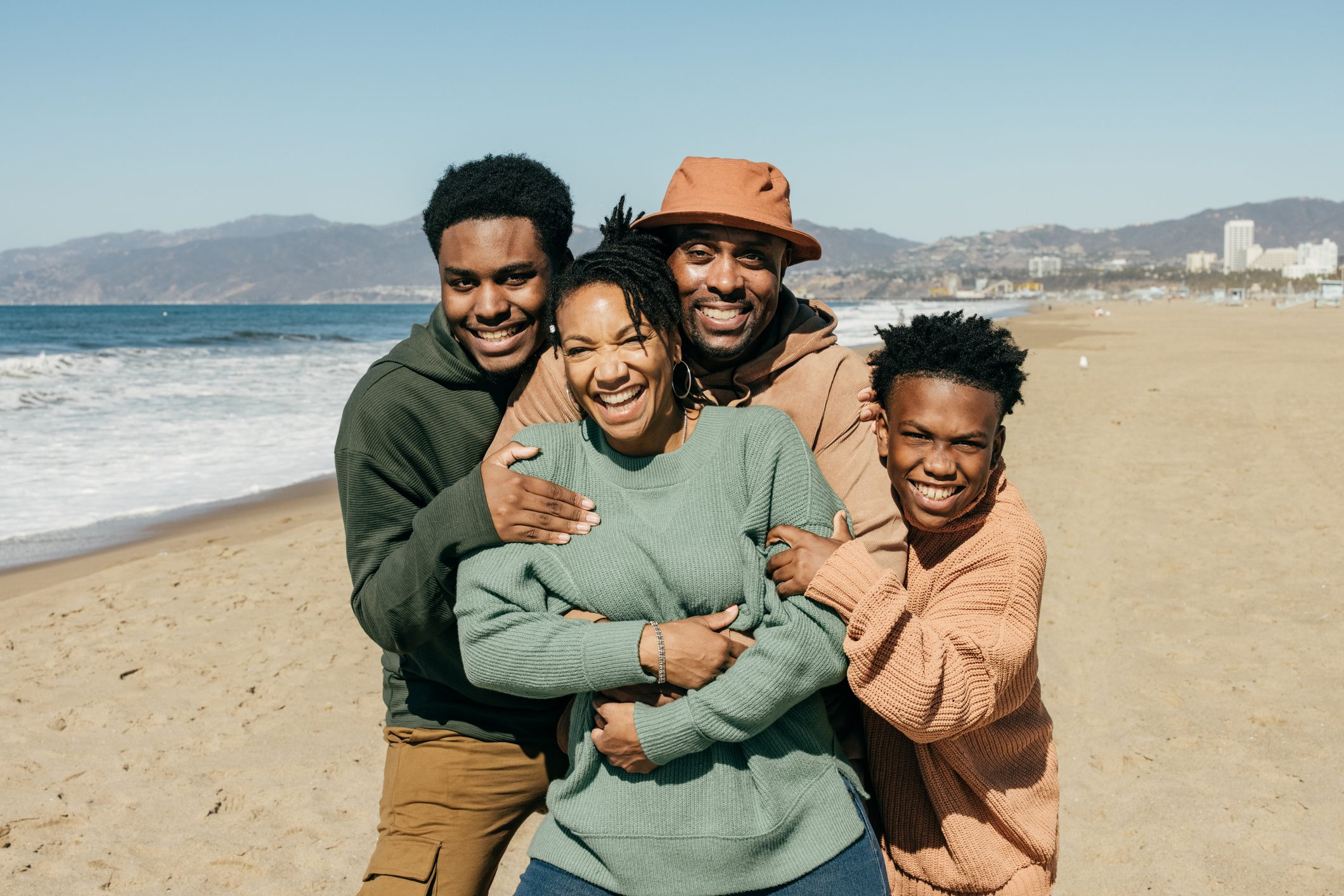 Family posing happily, embracing on a sandy beach with ocean waves; mountains and buildings visible in the background under a clear blue sky.