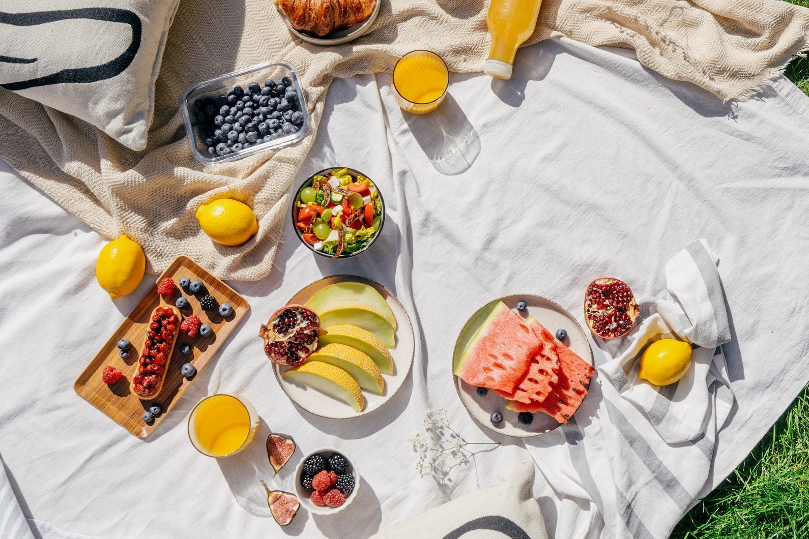 Fruits and pastries arranged on a picnic blanket, accented with dishes and orange juice, surrounded by grass on a sunny day.