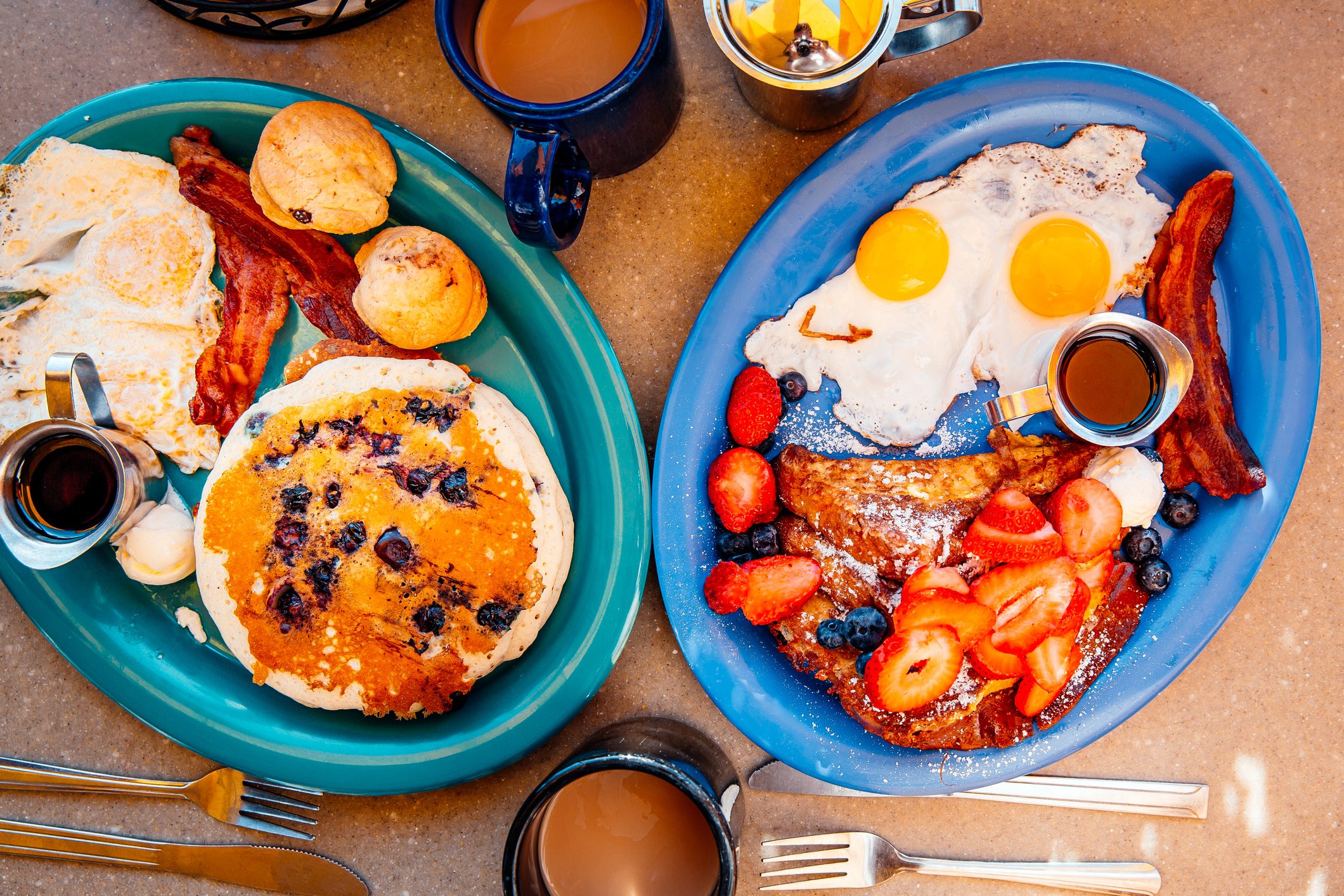 Two plates with breakfast items including eggs, bacon, pancakes, muffins, and fresh fruit. Coffee cups nearby on a wooden table.