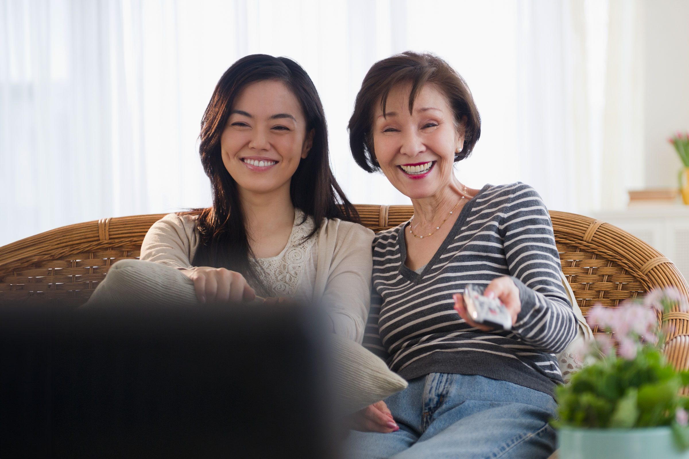 Two people sit smiling on a wicker couch, one holding a remote, with bright curtains and plants in the background.