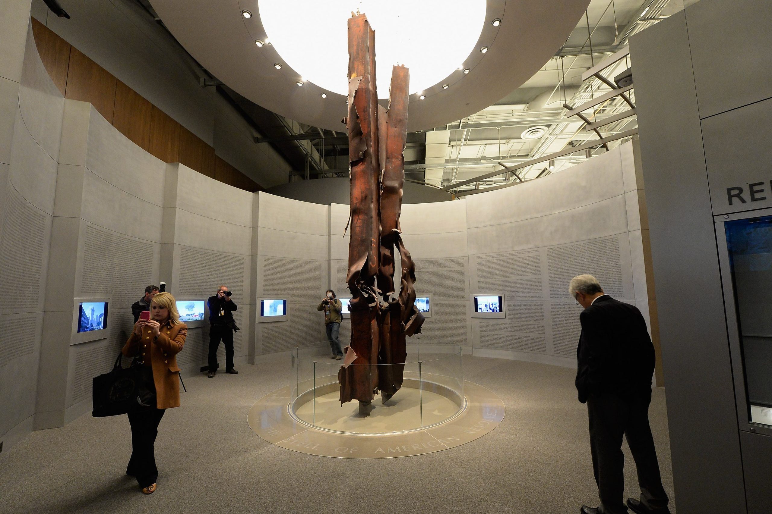 Twisted steel beams stand upright, surrounded by visitors photographing, within a museum environment with curved walls and display screens.