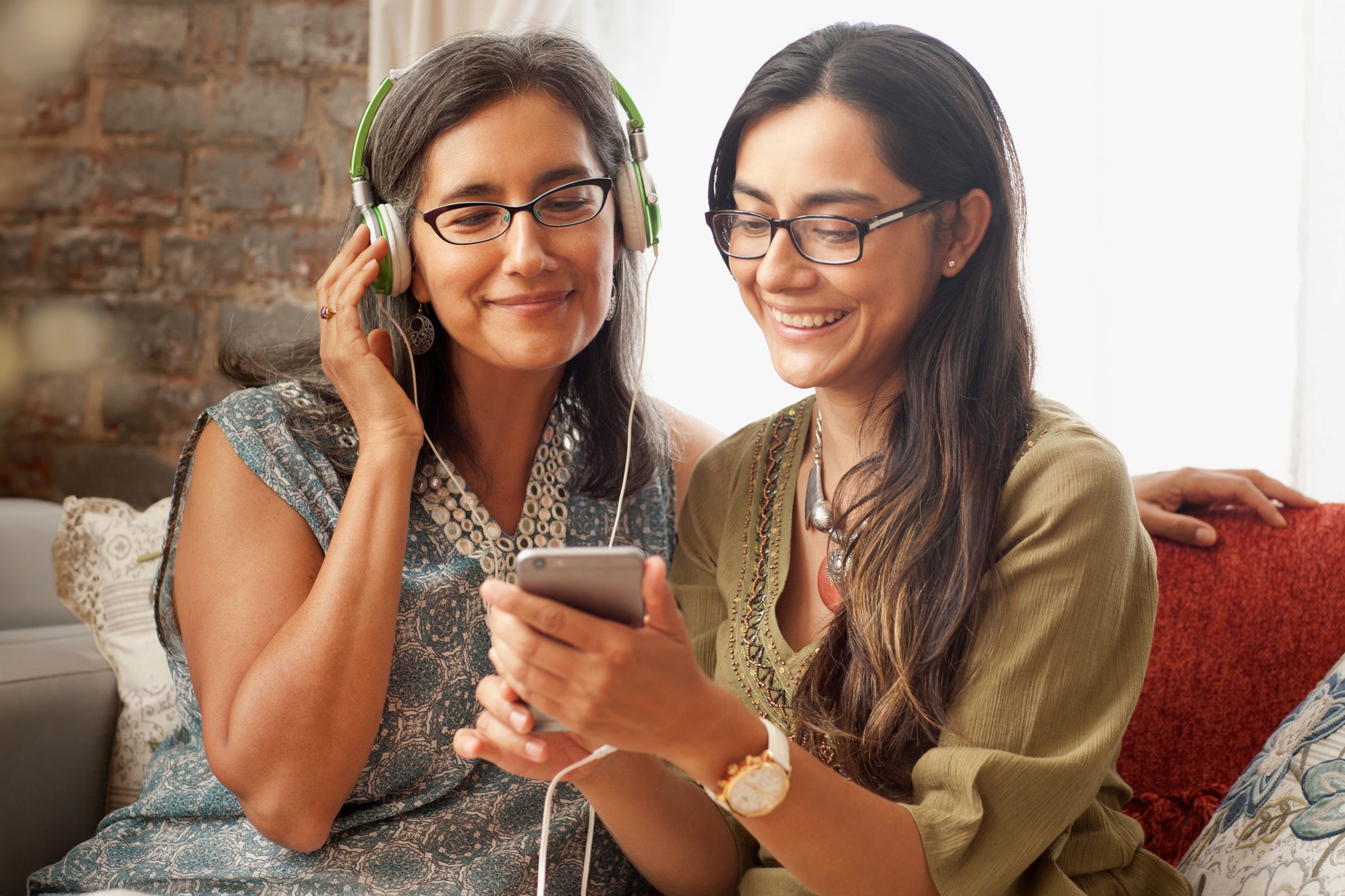 Two people share headphones, smiling at a phone, seated on a cozy sofa with a brick-wall backdrop.