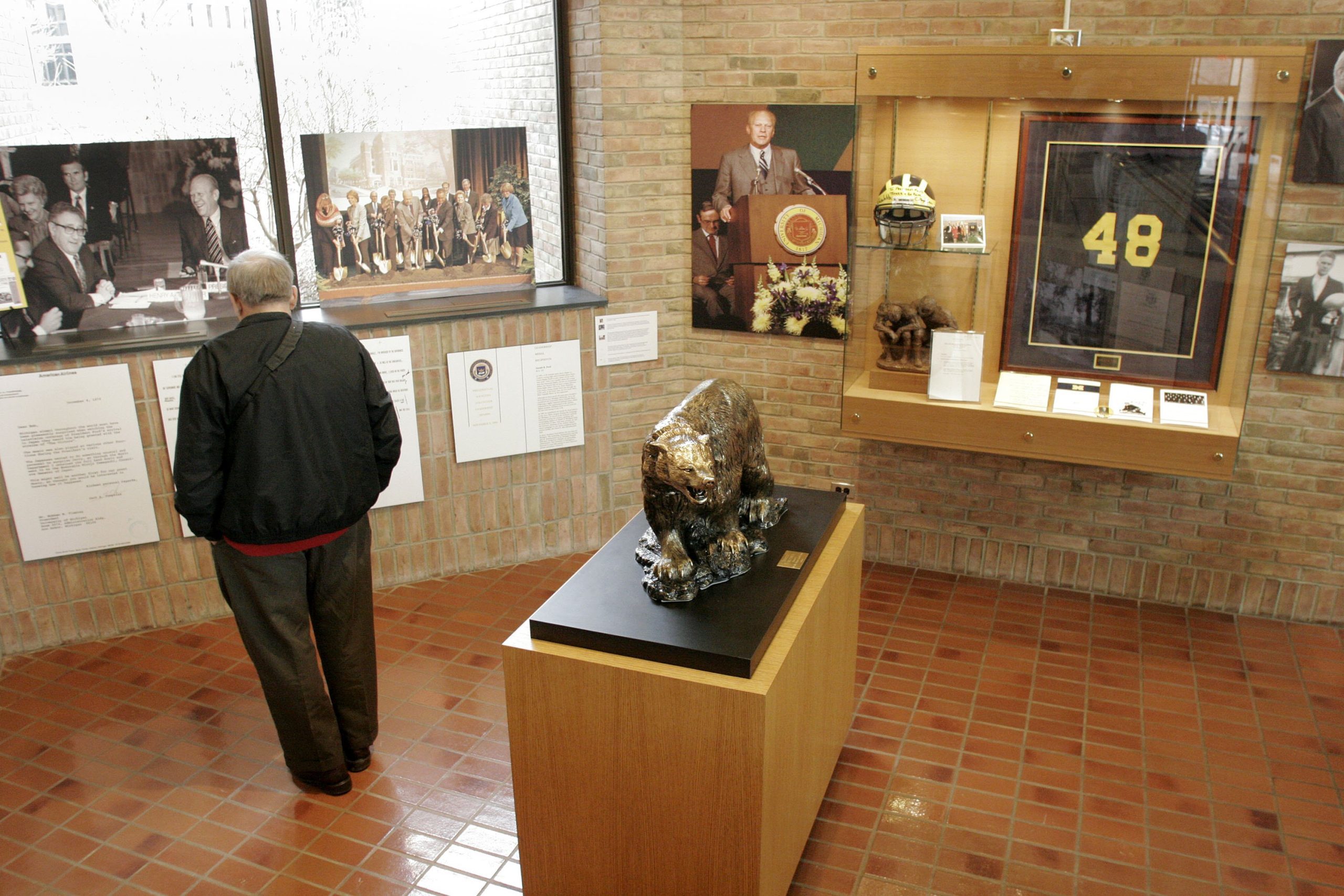 Statue of an animal displayed on a pedestal in a museum exhibit with a person observing nearby, surrounded by photographs and memorabilia on brick walls.