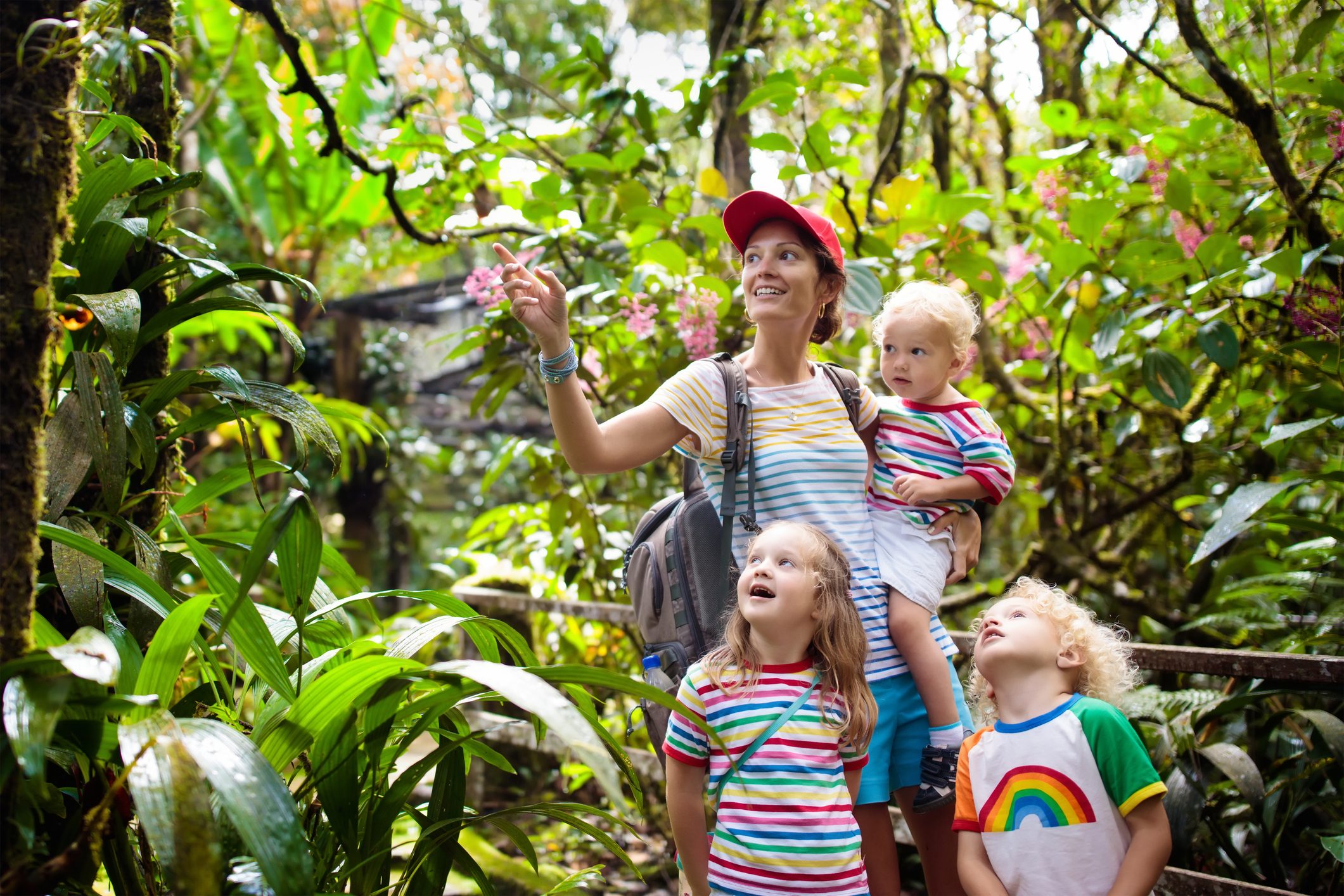 A woman points while exploring a lush, green jungle with three children wearing colorful striped shirts, surrounded by vibrant foliage.