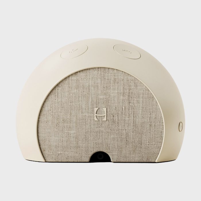 A rounded beige speaker is resting on a flat surface, featuring a fabric front with buttons on top in a neutral setting.