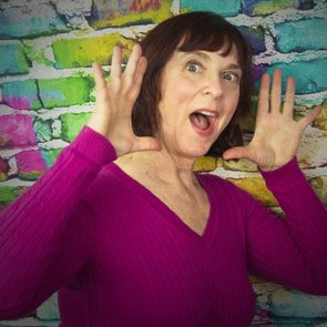 A person holds hands near face, smiling, against a colorful brick wall wearing a magenta top.