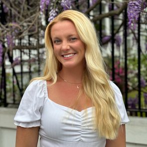 Woman smiling, standing outdoors; she's wearing a white top and necklace. Wisteria vines with purple flowers drape behind her against a black fence.