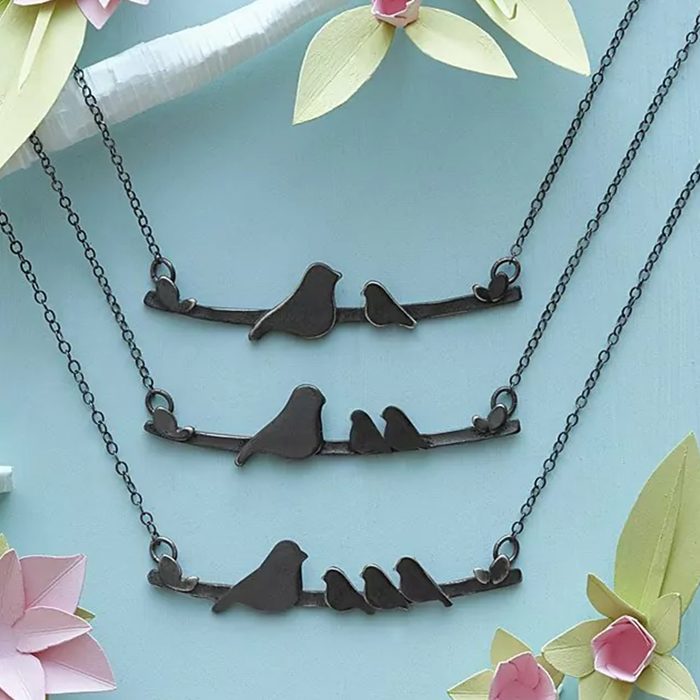 Necklaces depict bird silhouettes on branches, hanging against a soft blue background. Pastel paper flowers and leaves provide decorative accents.