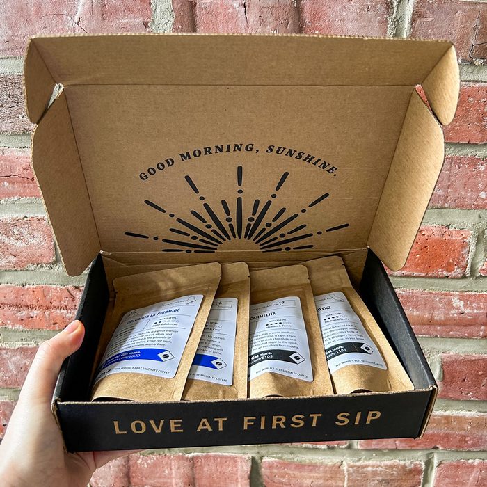 A hand holds a box containing four coffee packets, against a brick wall. Inside reads, 