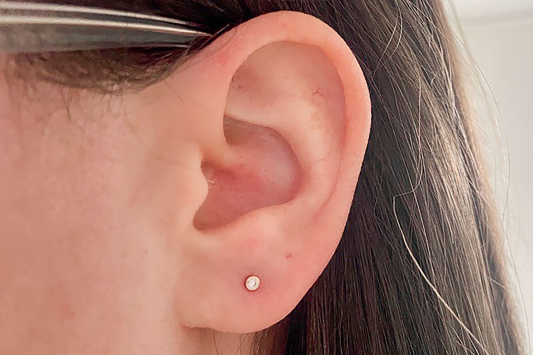 Earlobe displays a small, round stud earring; surrounded by dark hair in a well-lit setting.