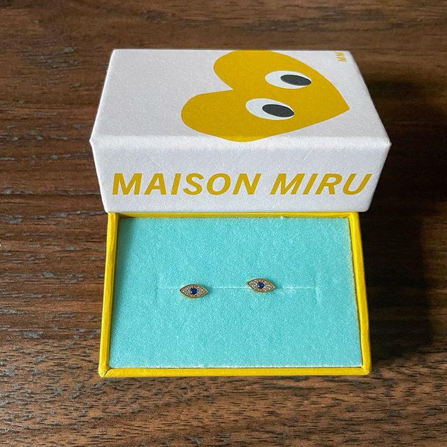 Earrings rest in an open jewelry box on a wooden table. The box lid displays text: 