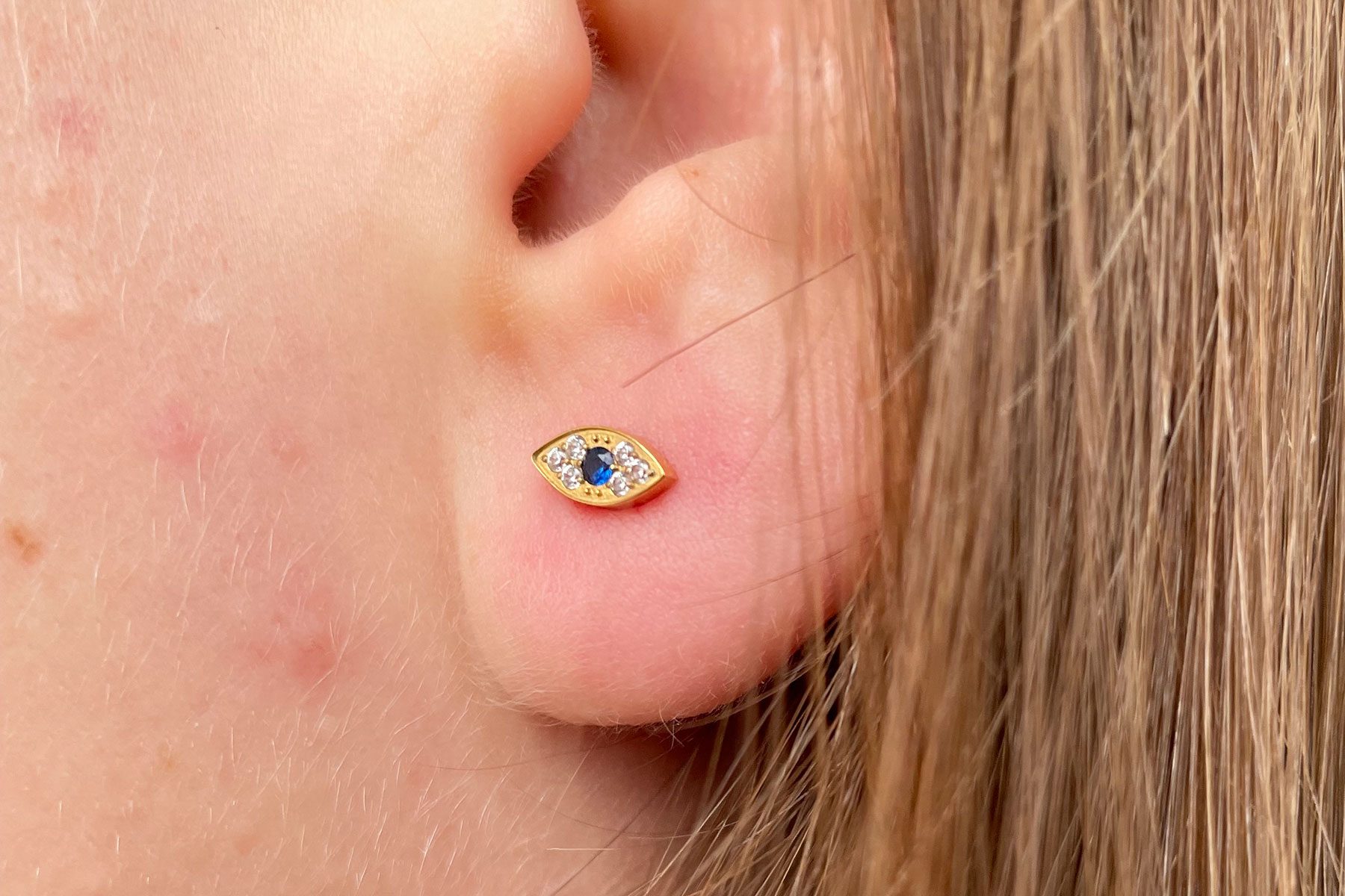 Gold stud earring sits in an ear; it features a central blue stone surrounded by small clear stones, with hair in the background.