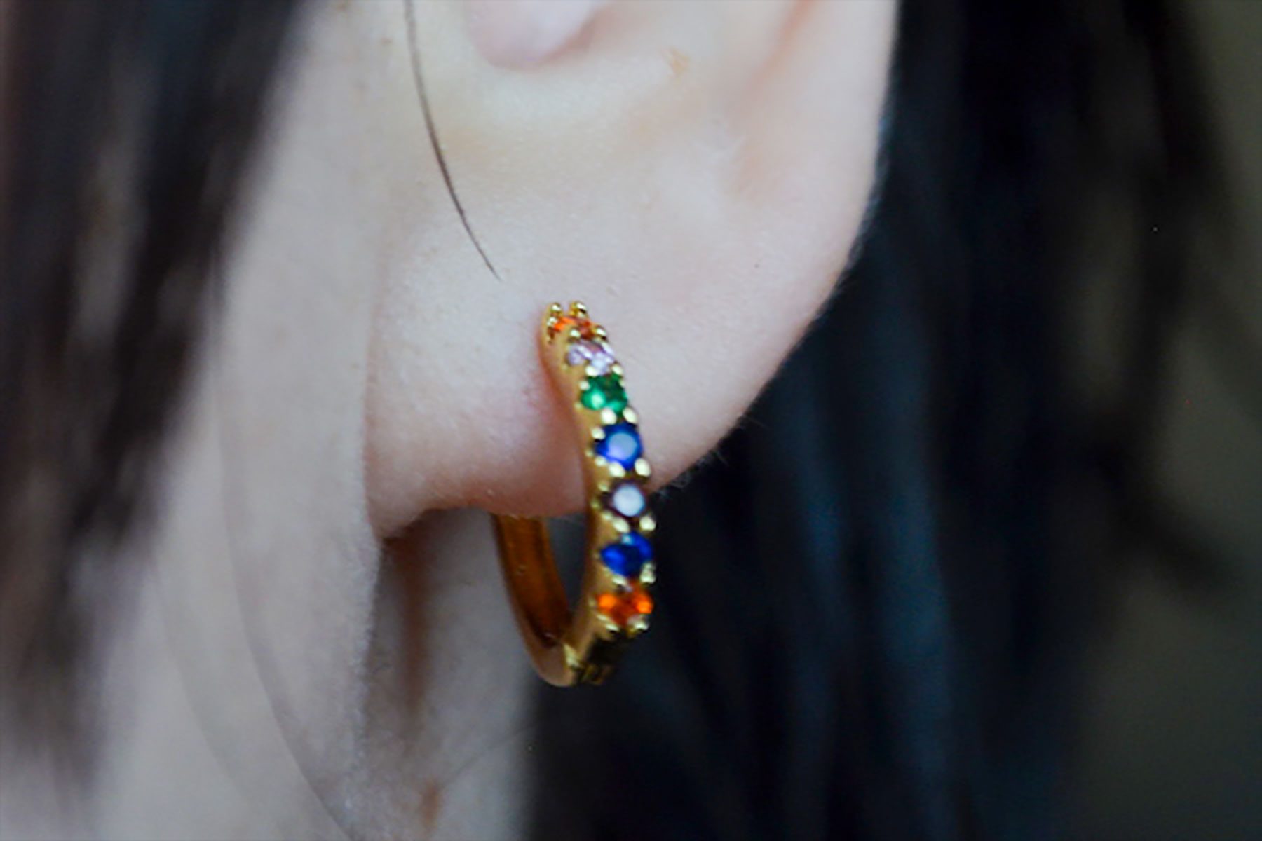 Colorful gemstone hoop earring adorns ear, surrounded by dark hair.