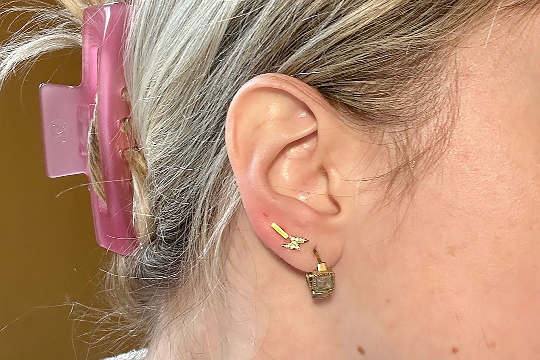 Ear with a lightning bolt earring, hair clipped back by a pink hair clip, against a plain background.