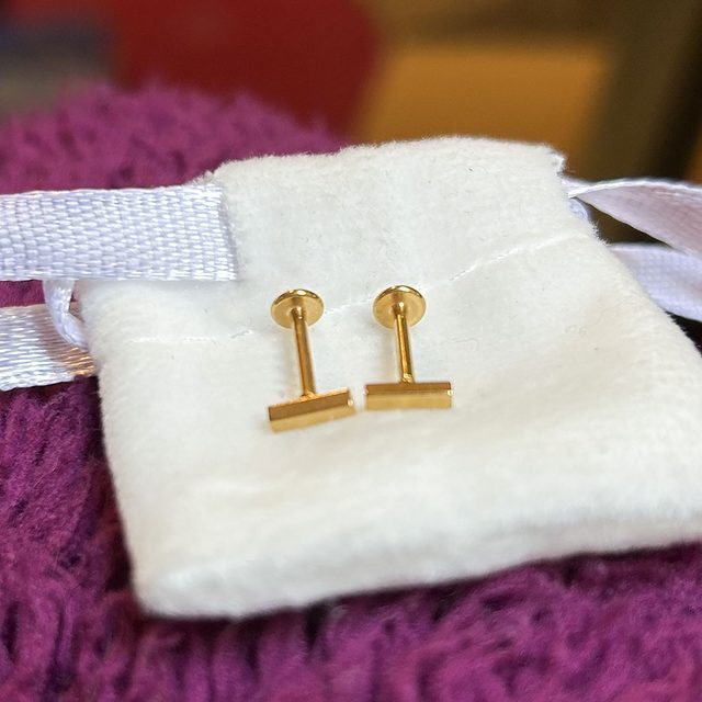 Gold barbell-style earrings sit on a soft white pouch, with a purple fabric in the background.