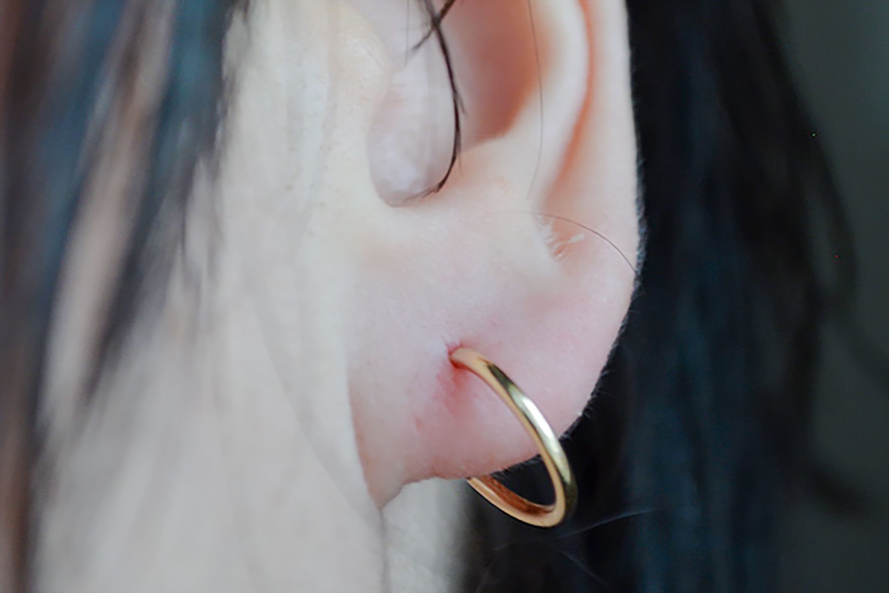 A gold hoop earring adorns an ear, partially obscured by dark hair strands in a dimly lit setting.