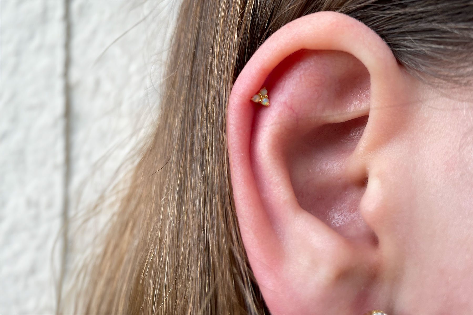 An ear displays a small opal piercing, surrounded by long, light brown hair in a textured, white-walled setting.