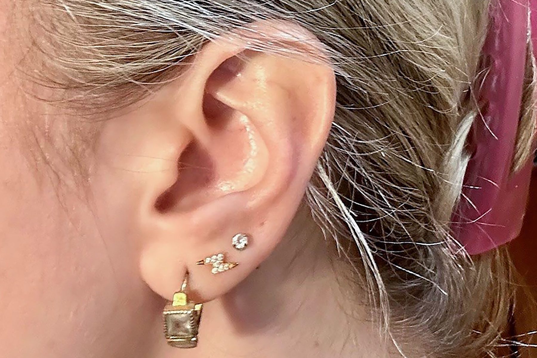 Ear adorned with three earrings, including a gold square hoop, diamond stud, and small arrow cluster. Blonde hair tied back with a pink band.