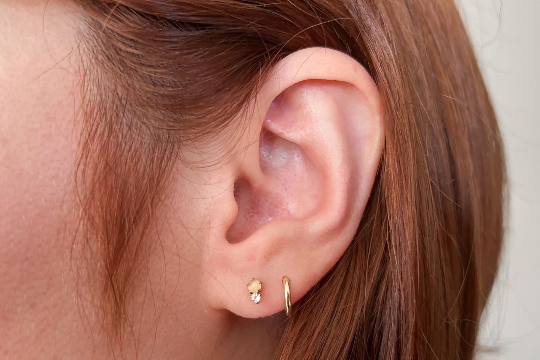 Ear adorned with two earrings, a small stud and a hoop; surrounded by brown hair.