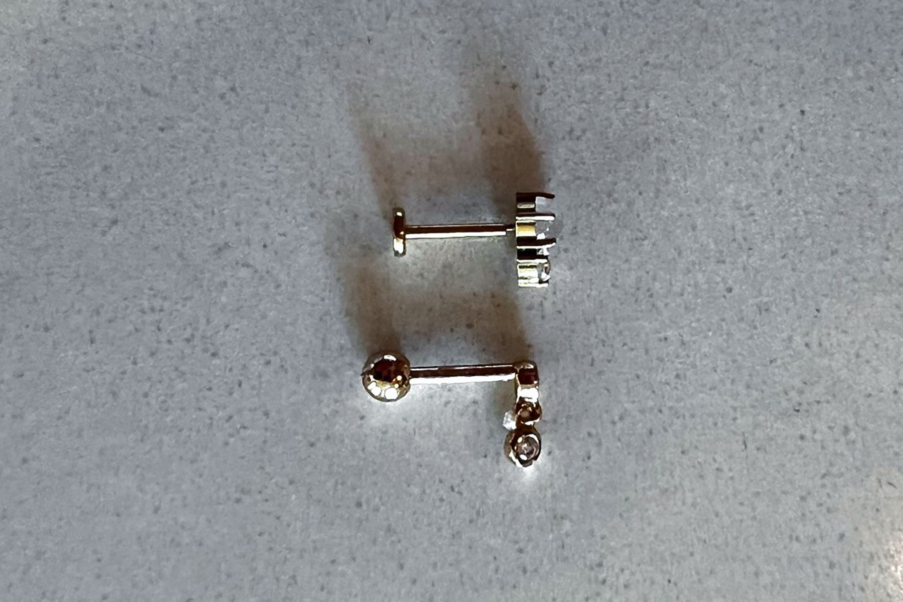 Two small, gold barbell piercings on a gray surface, reflecting light.