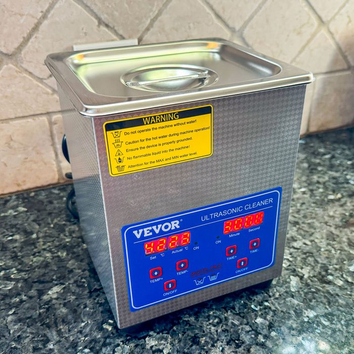 Ultrasonic cleaner sits idle on granite countertop, displaying temperature and time settings, surrounded by tiled backsplash.