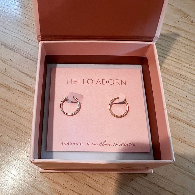 Earrings sit on a pink card inside an open box, resting on a wooden surface. The card reads 