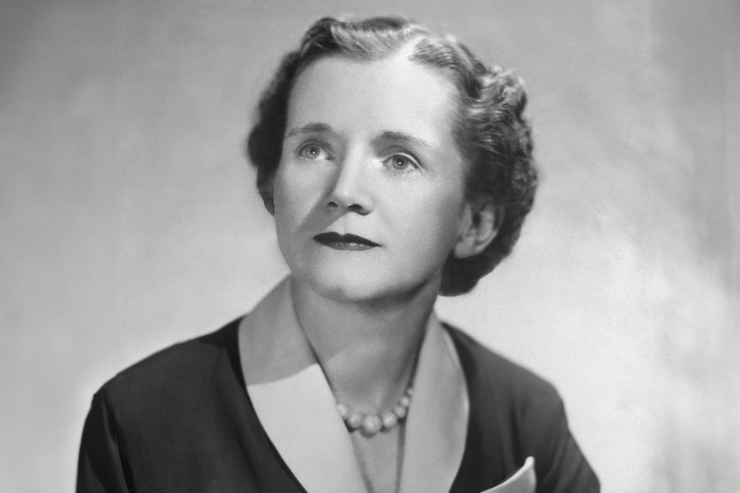 A woman in formal attire gazes upward, wearing a pearl necklace, set against a light, neutral background.
