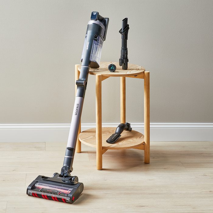 A cordless vacuum leans against a wooden shelf with accessories on a light wood floor in a minimally decorated room.