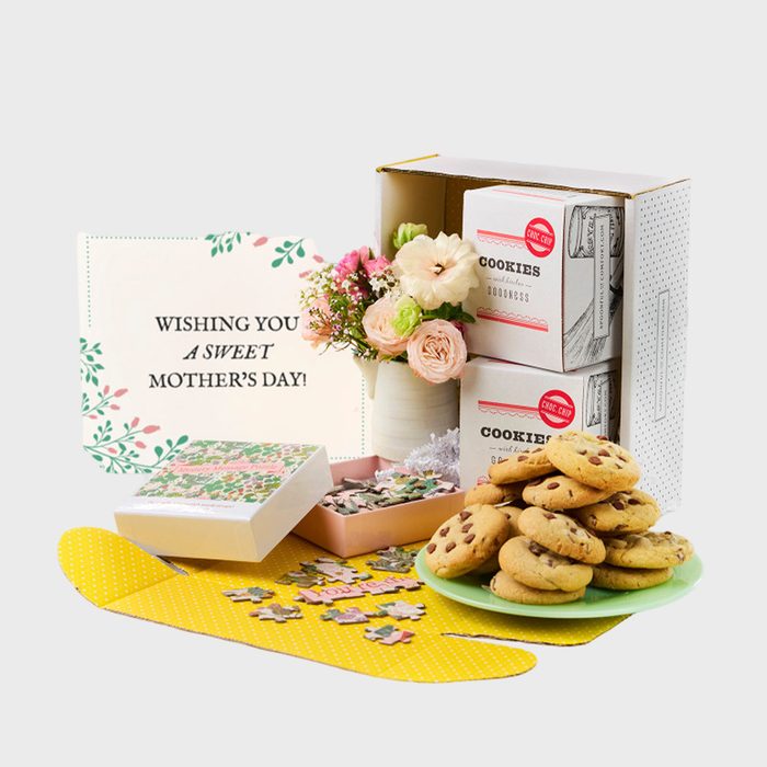 Cookies sit on a green plate, surrounded by flowers, a Mother's Day card, puzzle pieces, and cookie boxes in a gift set arrangement.