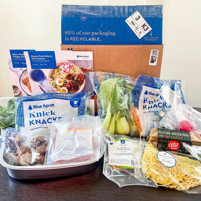 Food ingredients arranged. Various packaged items on a table, including vegetables, meats, and noodles. Blue Apron box visible in the background.