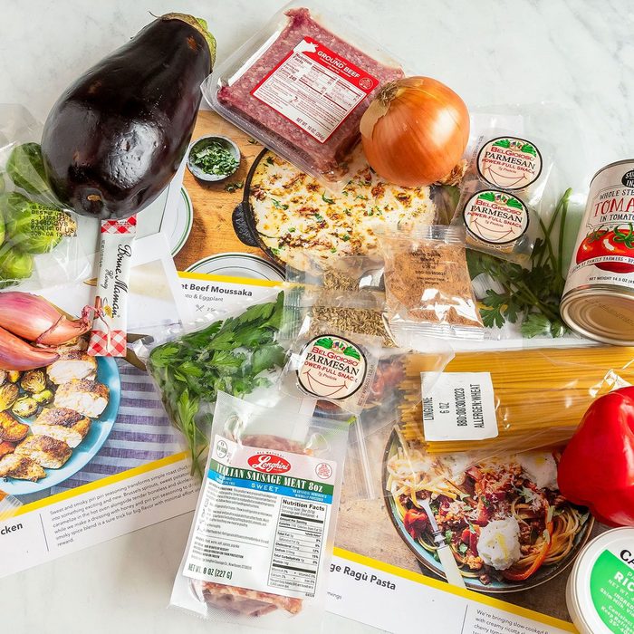 Ingredients and recipe cards are arranged on a table, including eggplant, onion, ground beef, cheese, pasta, spices, and canned tomatoes for cooking dishes.