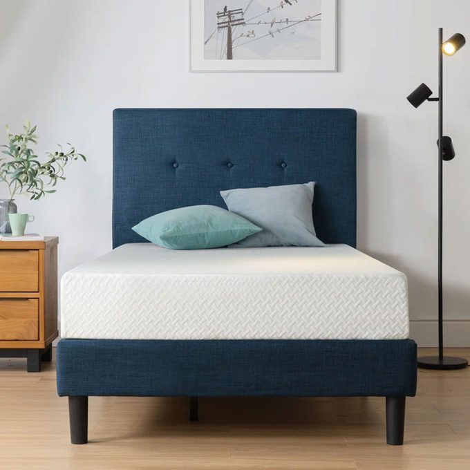 Bed stands in bedroom, displaying blue headboard. Two pillows rest atop a white mattress. Nearby, a wooden nightstand and a floor lamp.