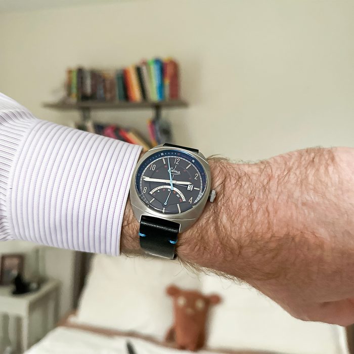 A wristwatch displays the time on an arm wearing a striped shirt, set in a bedroom with a blurred bookshelf and bed in the background.