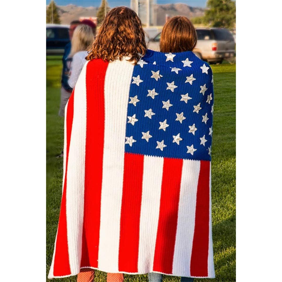 Two people wrapped in an American flag blanket stand on grass, with cars and trees in the background.