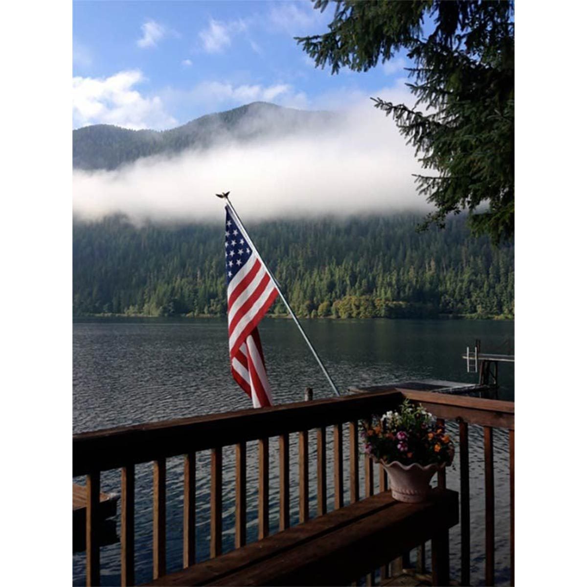 Flag waves on a deck overlooking a serene lake, surrounded by mountains and trees under a partly cloudy sky.