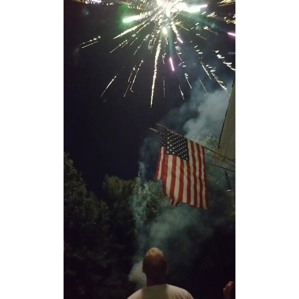 Fireworks burst overhead; a person watches beneath an American flag and trees, with smoke in the night sky.