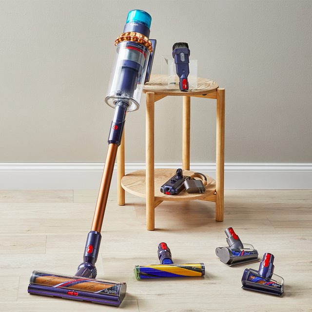 A vacuum cleaner leans against a wall; several attachments rest on a wooden table and floor in a minimalist room.